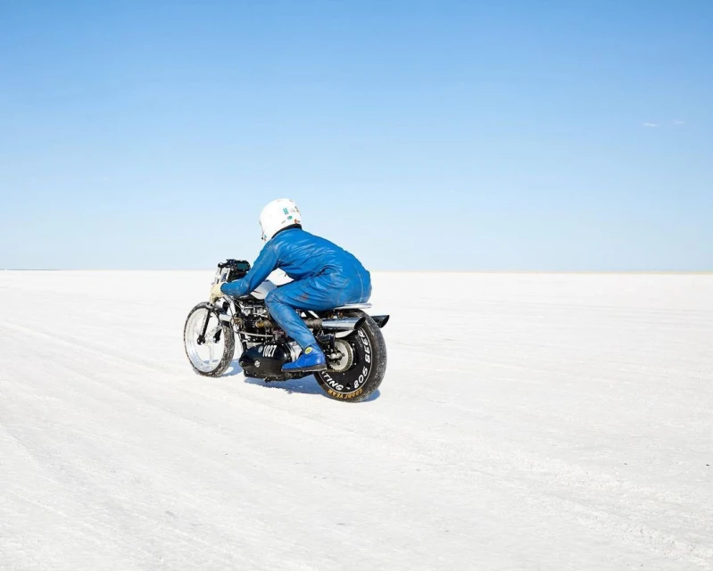 Motorcycle racer in blue suit speeds across a white salt flat under a clear blue sky. Action shot from the rear.