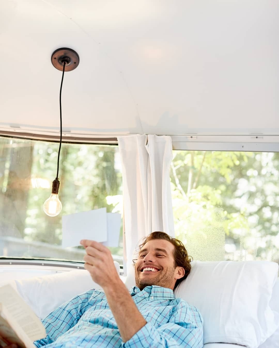 Man relaxing in camper with book and cards, smiling, under a hanging lightbulb, surrounded by white walls and forest view.
