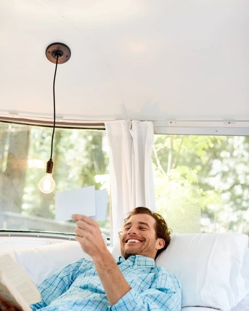 Man relaxing in camper with book and cards, smiling, under a hanging lightbulb, surrounded by white walls and forest view.