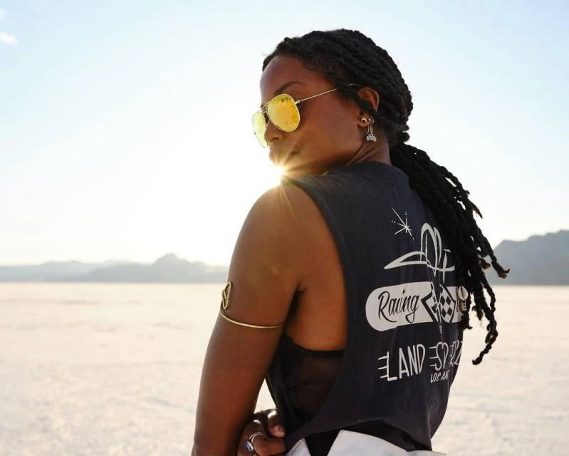 Adult female in sunglasses and muscle shirt poses in the sun against a desert backdrop, wearing jewelry and facing away from the camera.