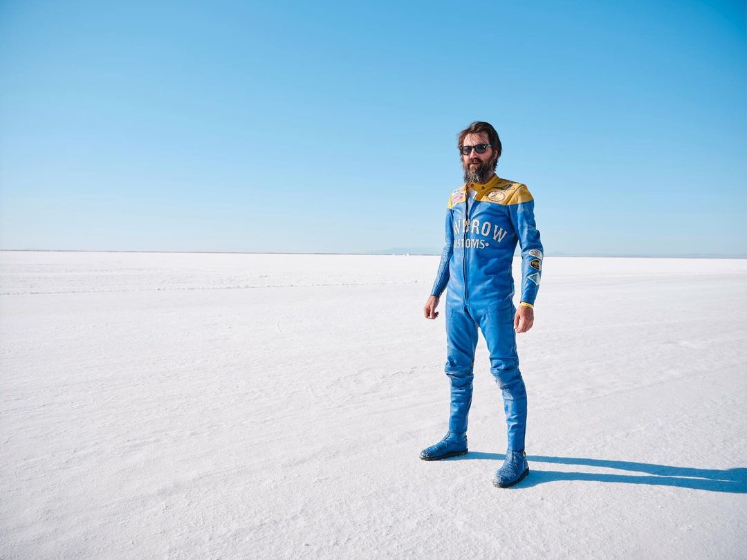 Adult male in blue racing suit with gold trim standing in desert salt flats under bright daylight, sunglasses, beard, full shot.
