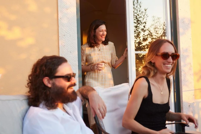 Three smiling people gathered outdoors in warm evening light, with a woman standing in a doorway holding a glass.