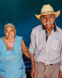 Elderly man in a straw cowboy hat and elderly woman in a blue plaid apron standing side by side in front of a textured blue wall.