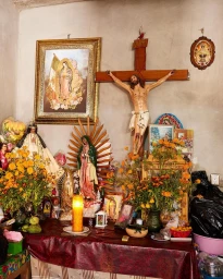 Religious altar decorated with statues of Our Lady of Guadalupe, a wooden crucifix, framed portraits, and orange flowers by David Martinez