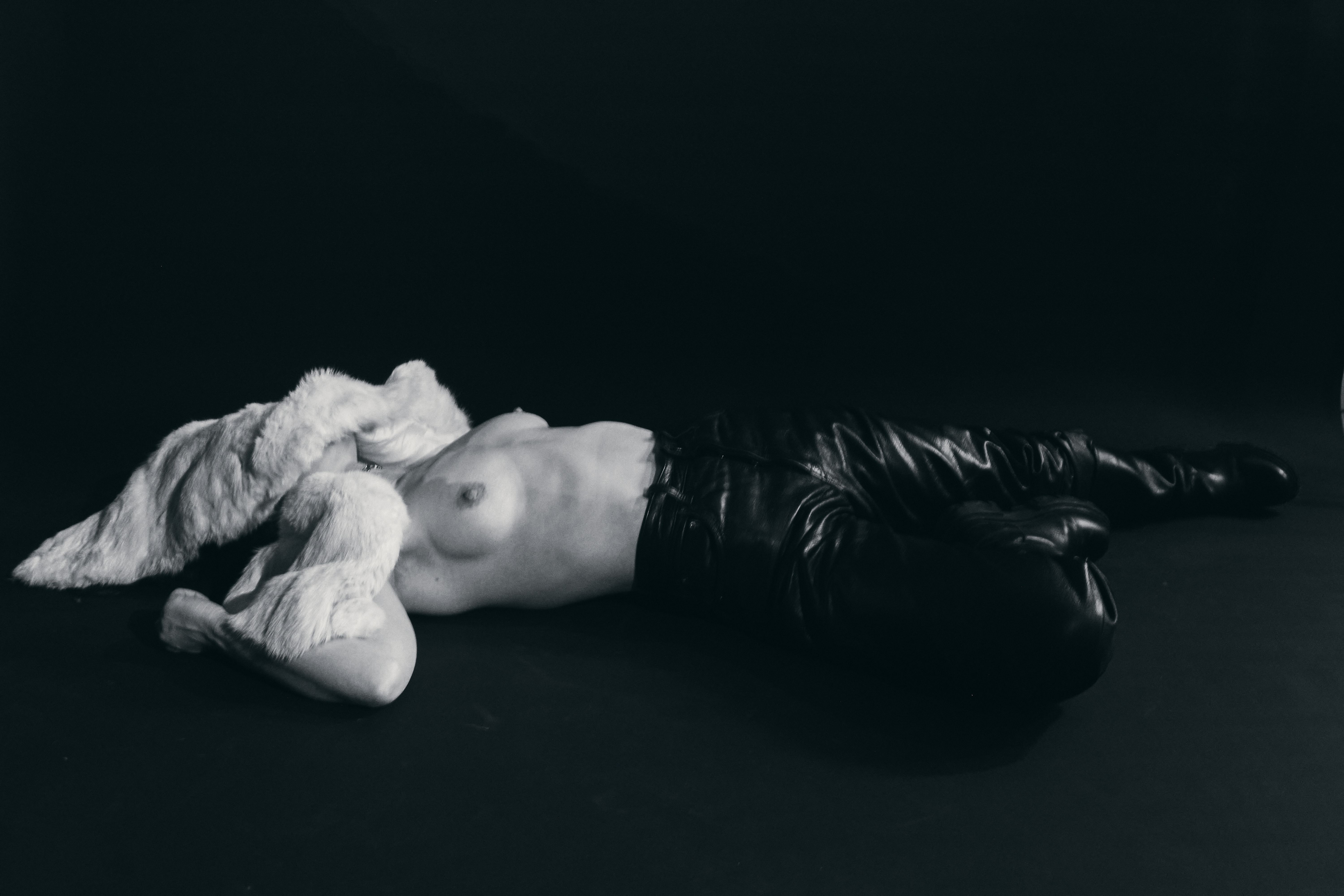 Artistic black and white portrait of a nude female torso with white furry object, set against a dark monochromatic background.