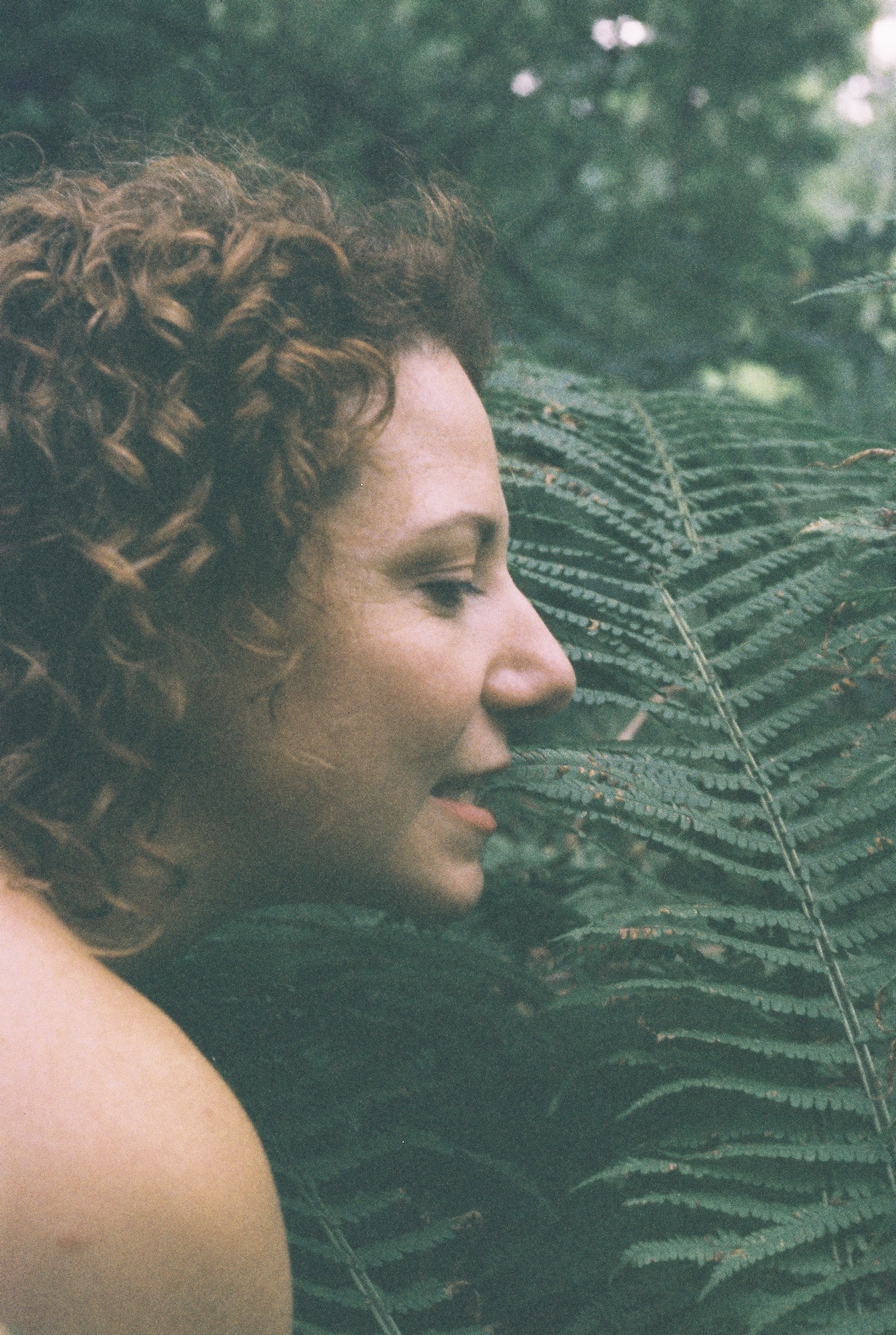 Adult female portrait in profile, outdoors, with curly auburn hair. Woman is positioned next to a large fern, facing to the right.