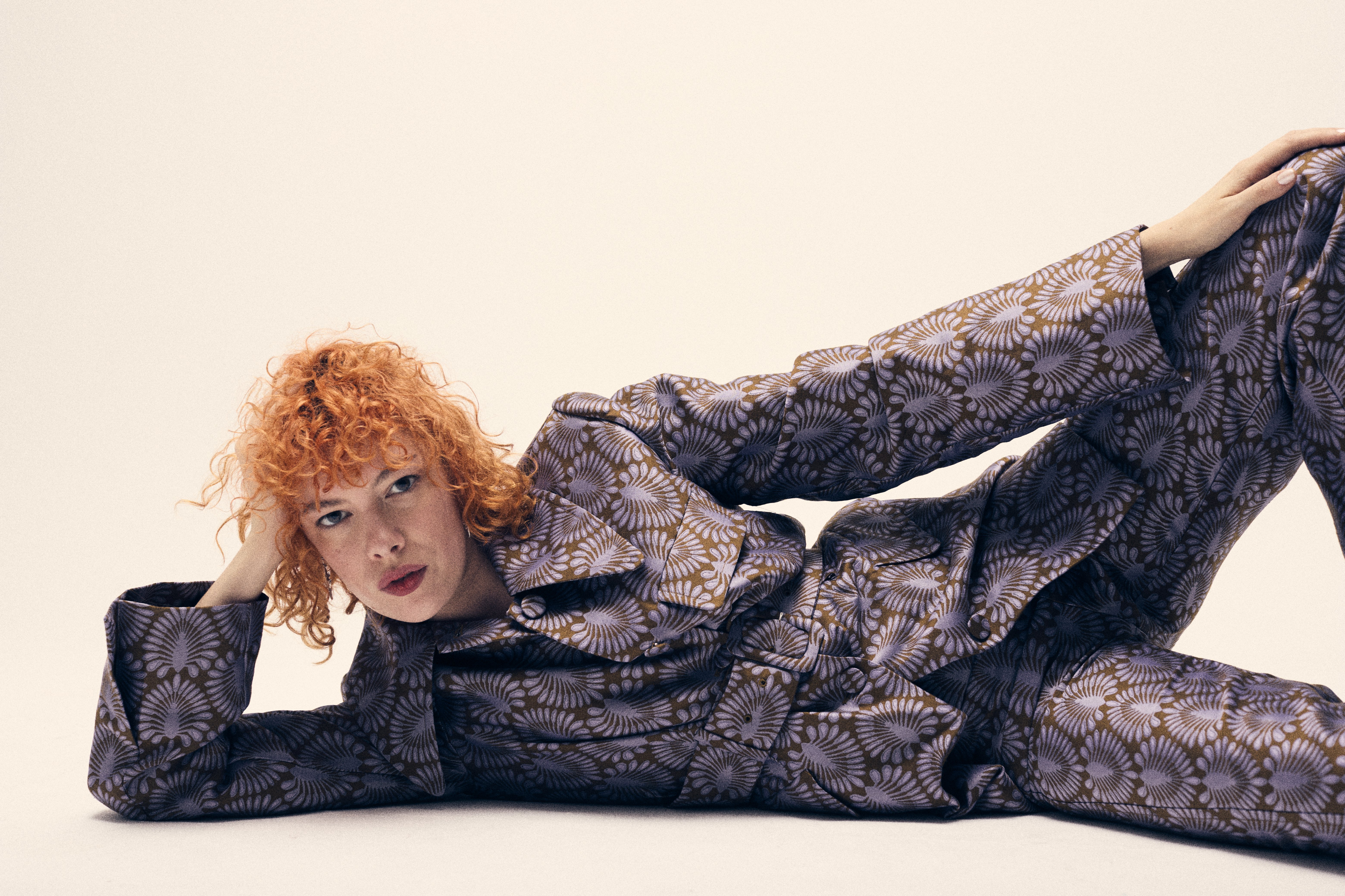 Woman with red curly hair, wearing patterned lilac suit, poses on the floor in a studio setting, looking at the camera with a neutral expression.
