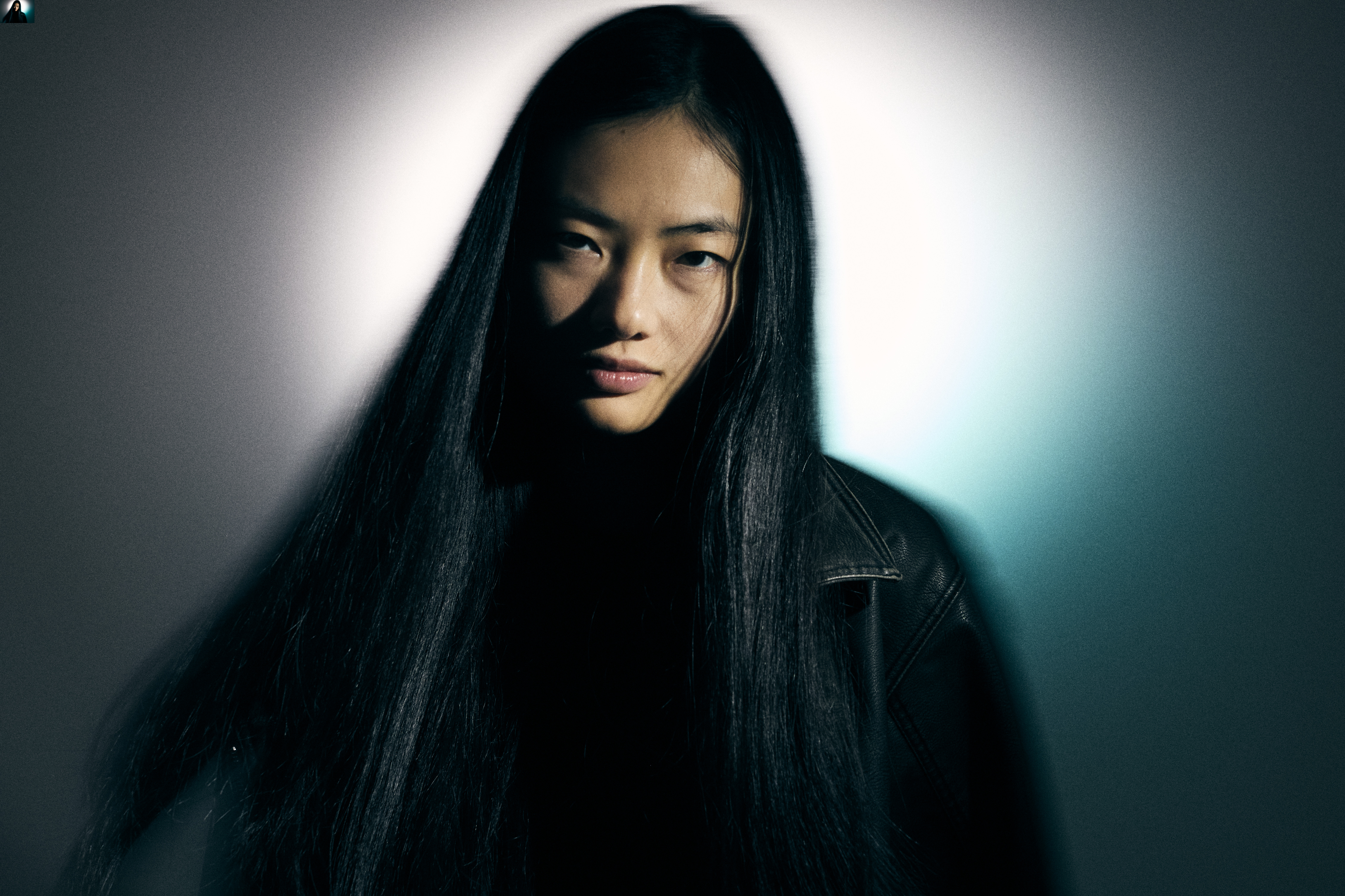 Portrait of an Asian woman with long dark hair wearing a black leather jacket against a two-tone light blue and white background.