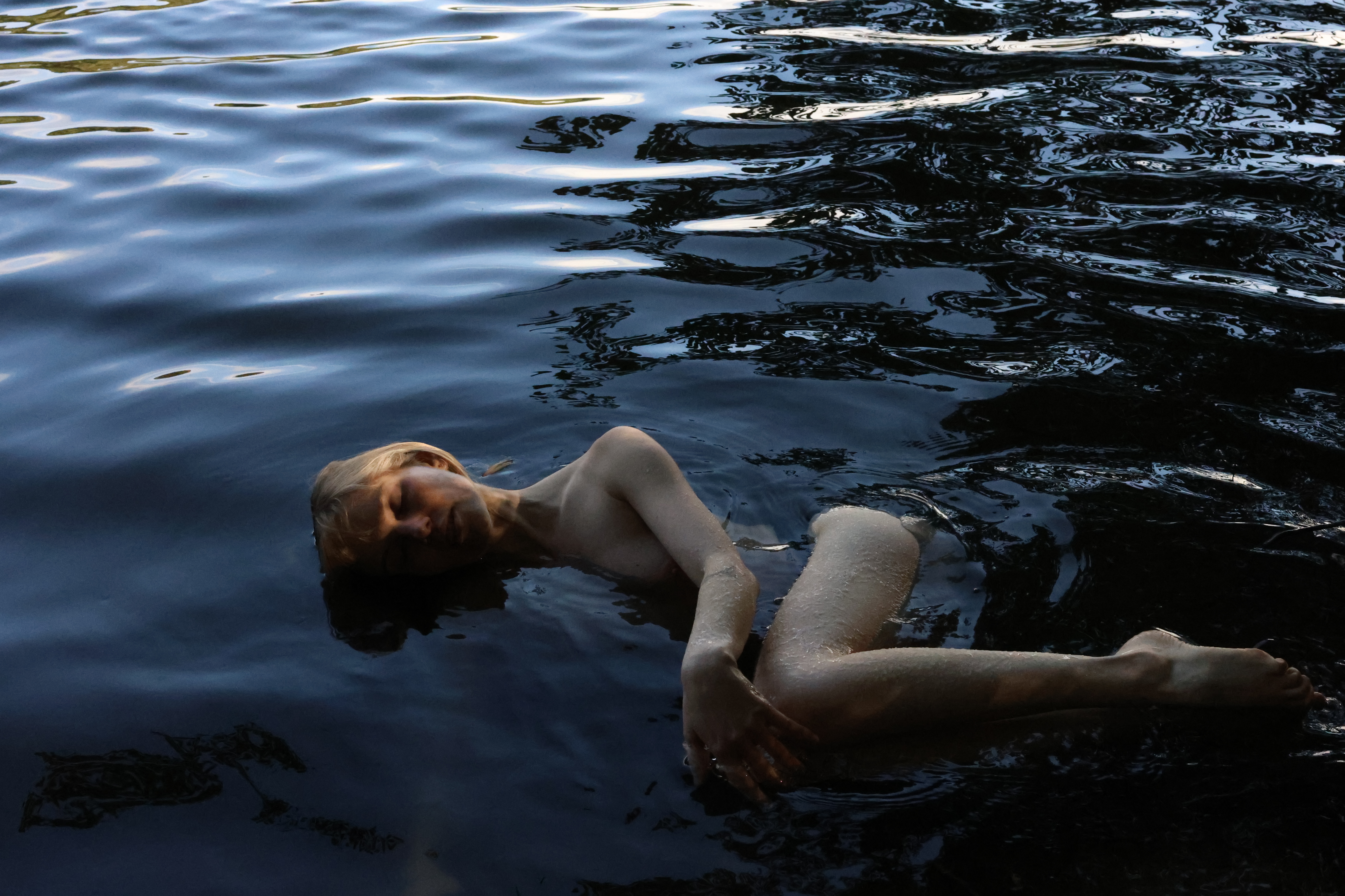Woman with fair hair reclines in dark, rippled water, creating a serene and somewhat surreal image with natural lighting and reflections.