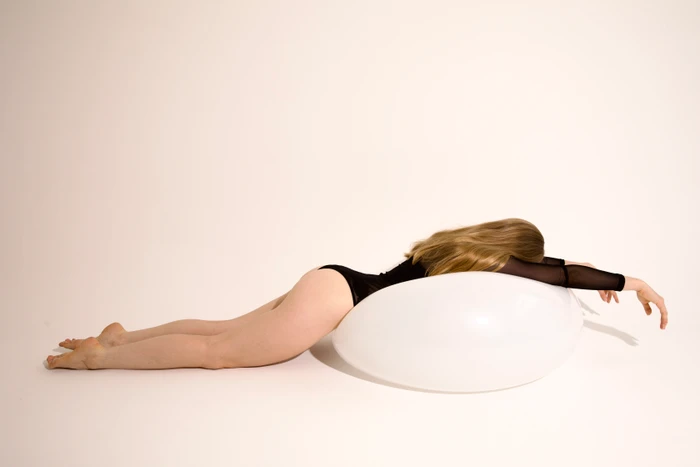 A woman in a black bodysuit lies prone across a large white inflated sphere on a seamless studio background.