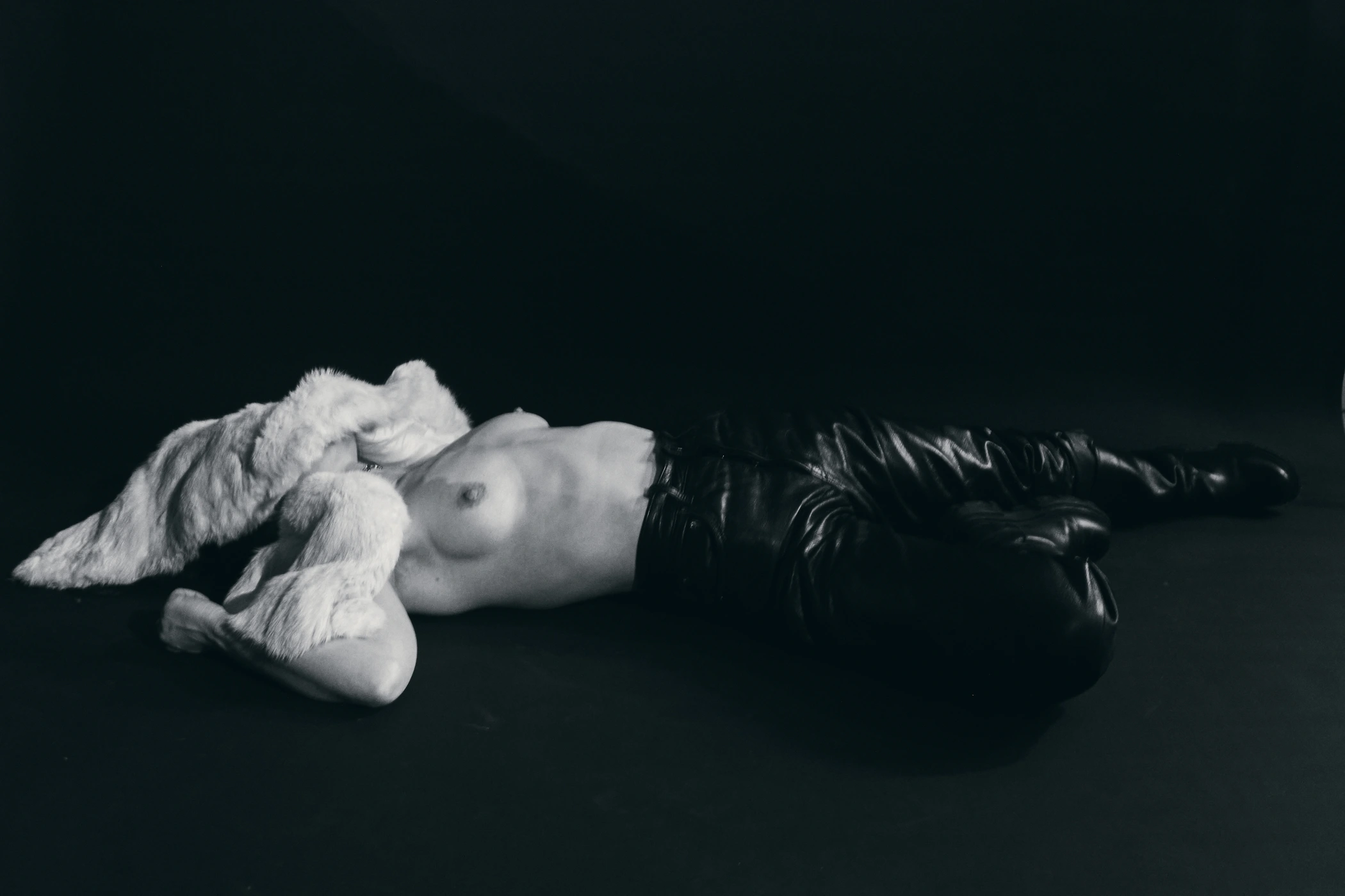 Model lying on a black floor wearing black leather pants and a white textured fur stole draped over her face.