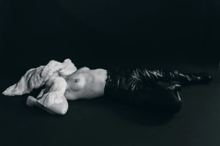Model lying on a black floor wearing black leather pants and a white textured fur stole draped over her face.