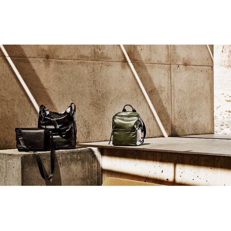 Three bags are displayed on a concrete surface with shadows. There is a small backpack and two tote bags, styled for fashion.