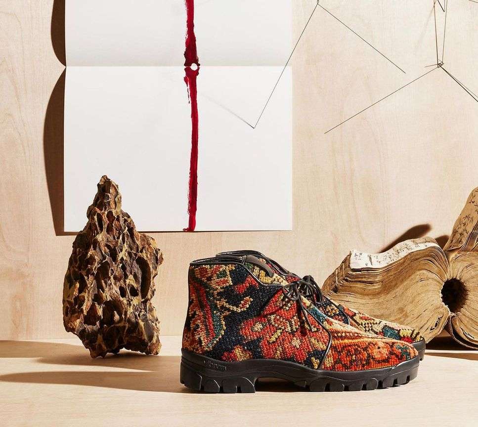 Textured ankle boots with graphic pattern, set against geometric paper art, aged book, and a porous rock formation on a wood surface.