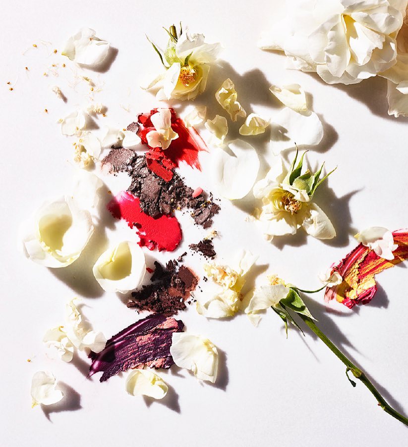 Cosmetics with lipstick, eyeshadow, and white rose petals scattered on a white surface in a beauty product flat lay.