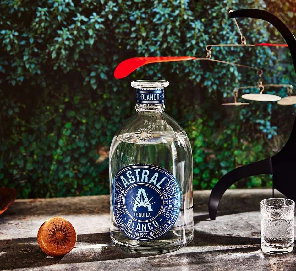 Astral Tequila Blanco bottle with a glass, and a wooden object against a leafy background. Tequila still life product shot.
