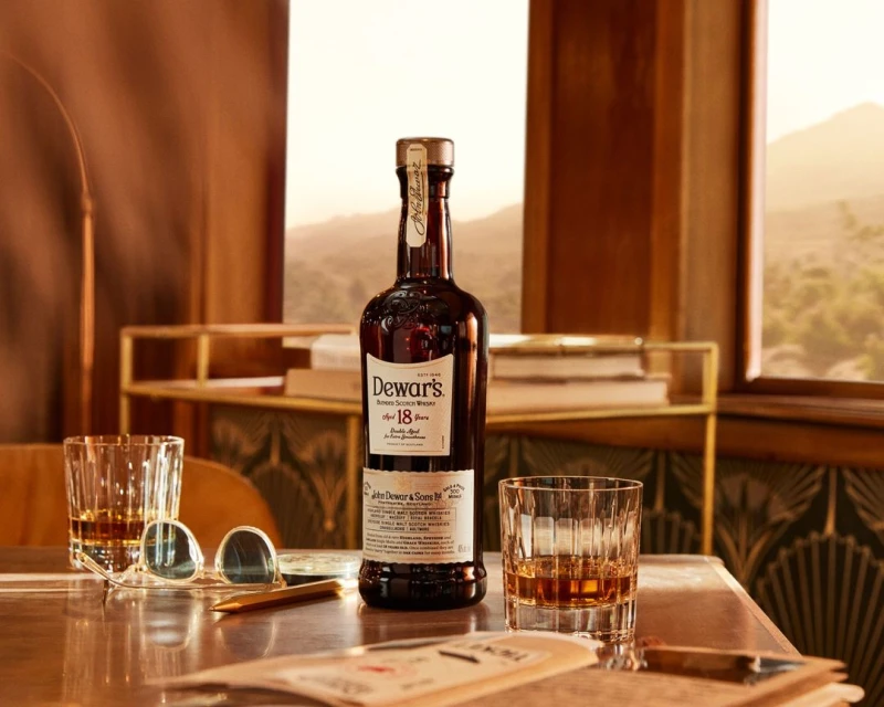 Dewar's 18-year-old whisky bottle on a table with glasses, against a scenic backdrop, highlighting the product's aged quality and setting.