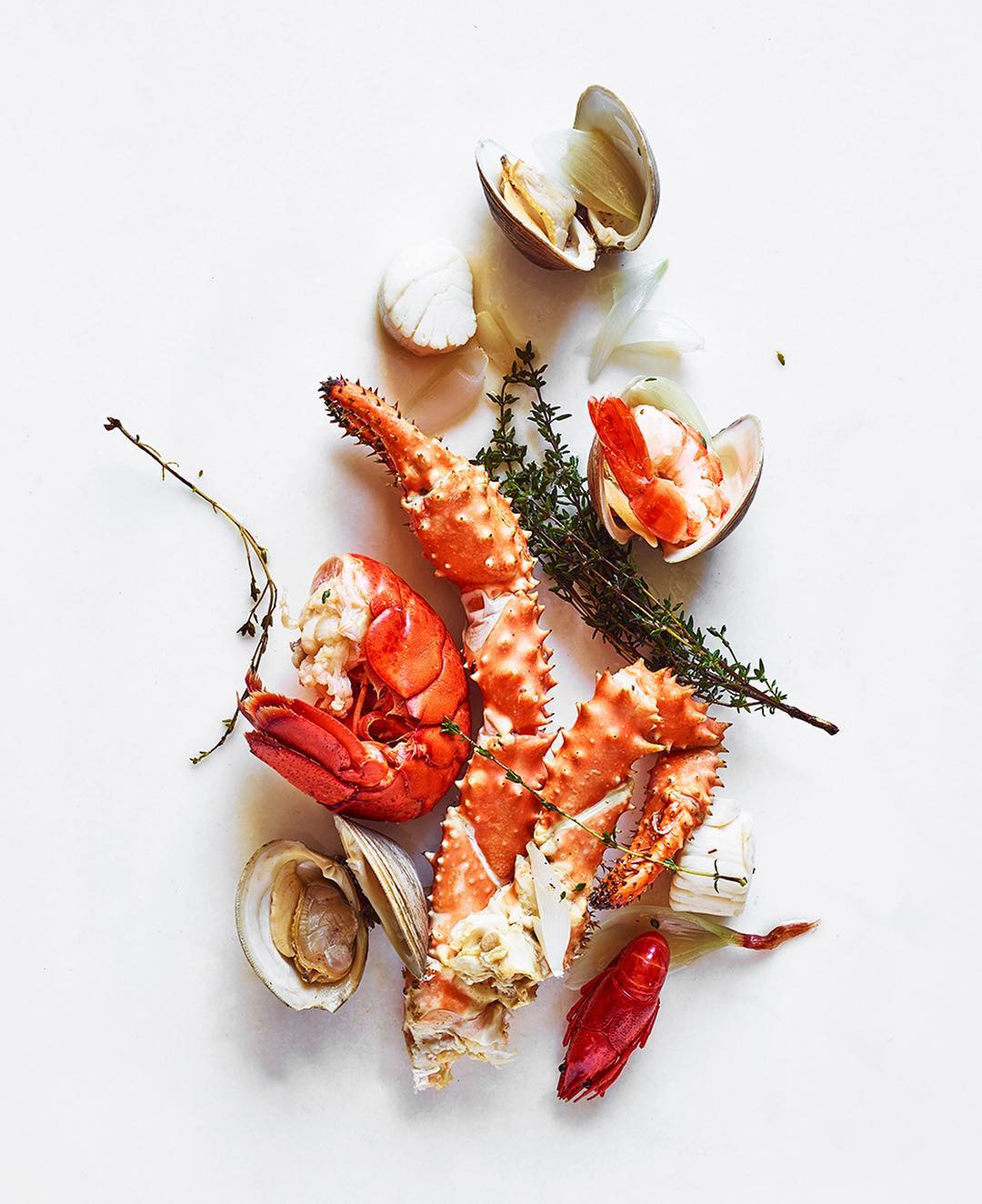 Overhead shot of an assortment of seafood including king crab legs, shrimp, clams, and crawfish, arranged on a white background with herbs.