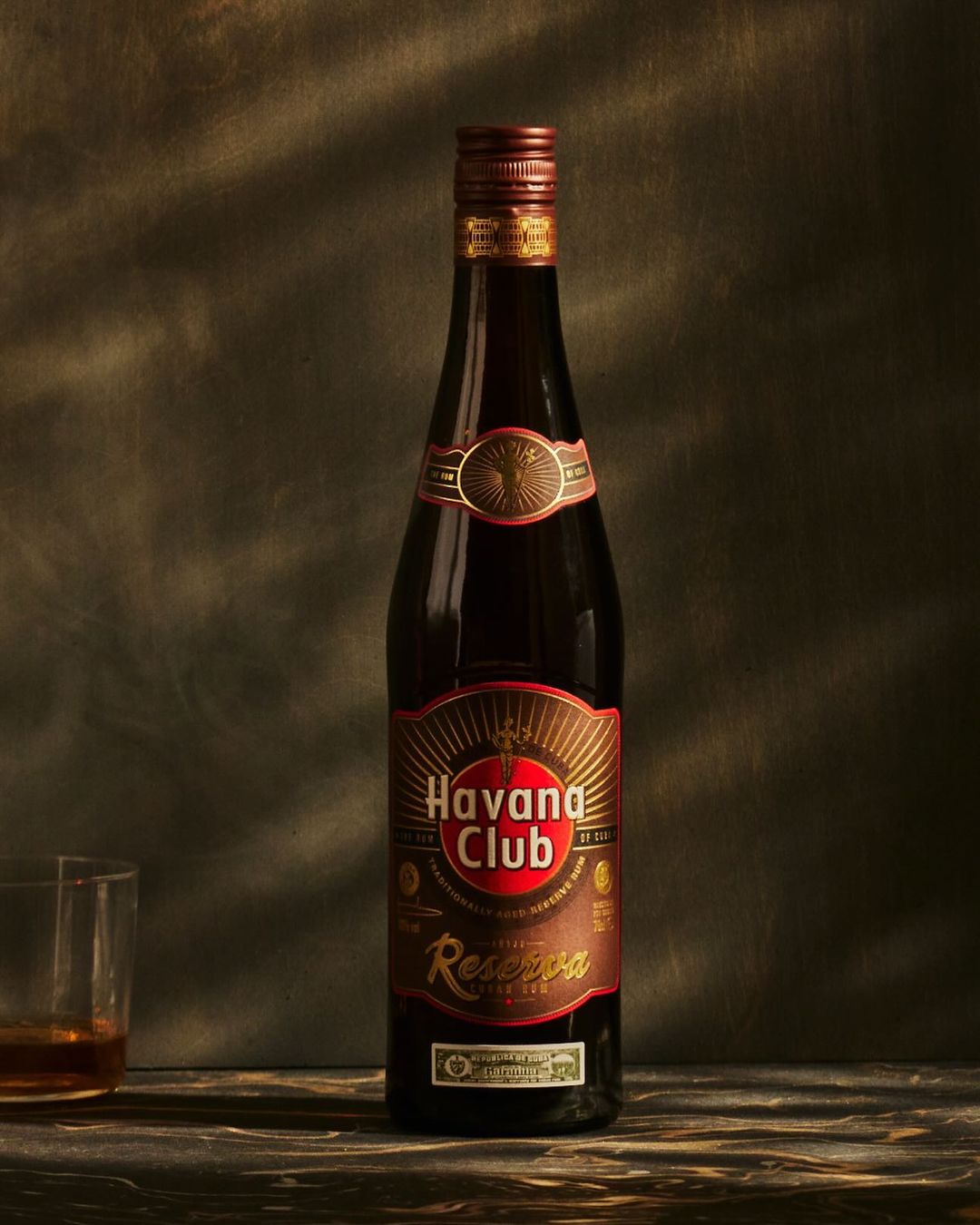 Bottle of Havana Club Reserva rum on a dark surface with glass of rum, against a textured background, warm lighting, close-up.