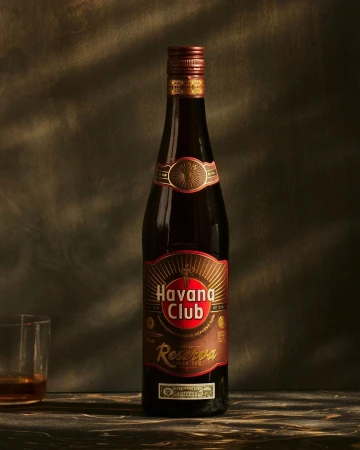 Bottle of Havana Club Reserva rum on a dark surface with glass of rum, against a textured background, warm lighting, close-up.