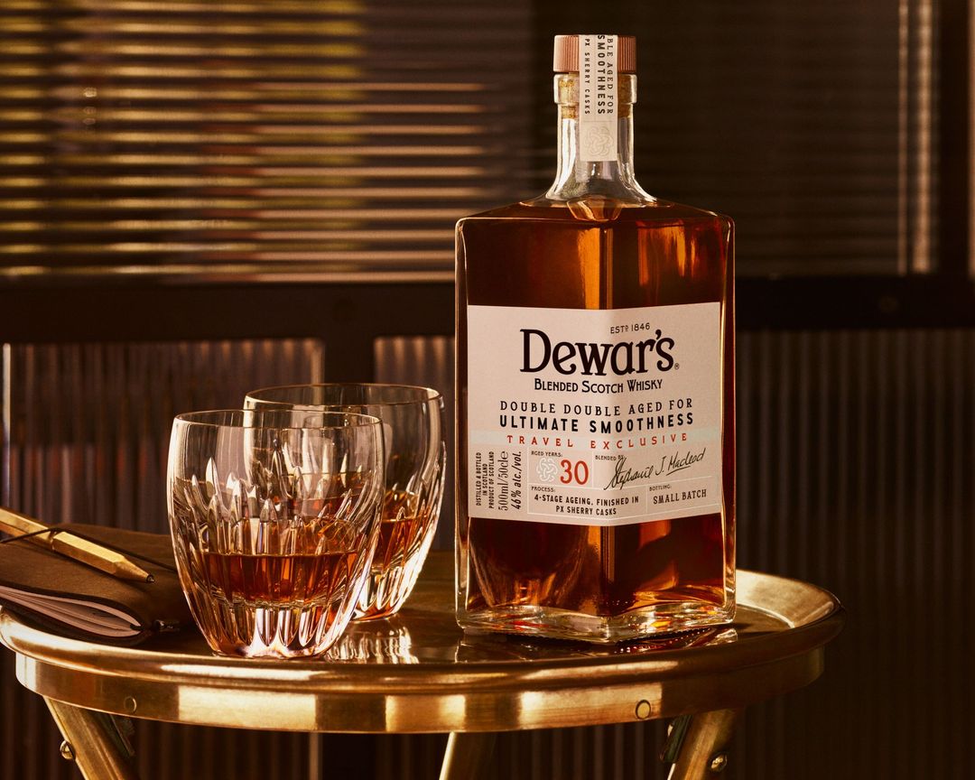 Dewar's blended Scotch whisky bottle with two crystal glasses filled with whisky on a gold side table in a dimly lit room.