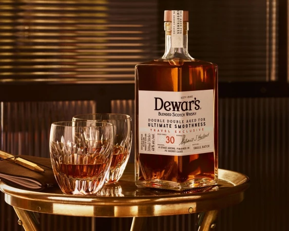 Dewar's blended Scotch whisky bottle with two crystal glasses filled with whisky on a gold side table in a dimly lit room.