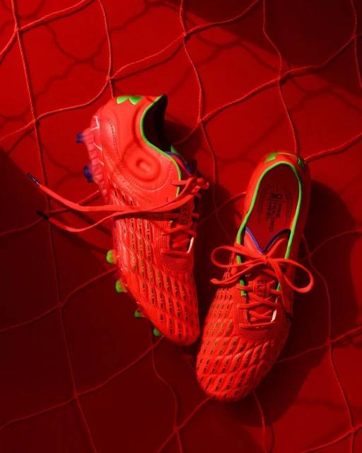 Two red and green soccer shoes are set on a matching red net, studio shot with bright, directional lighting and vibrant color grading.