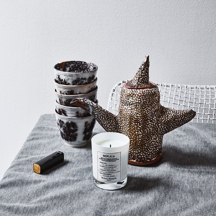 Still life featuring a stack of ceramic cups, a decorative ceramic teapot, and a REPLICA scented candle on a linen-covered table.