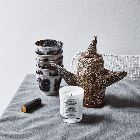 Still life featuring a stack of ceramic cups, a decorative ceramic teapot, and a REPLICA scented candle on a linen-covered table.
