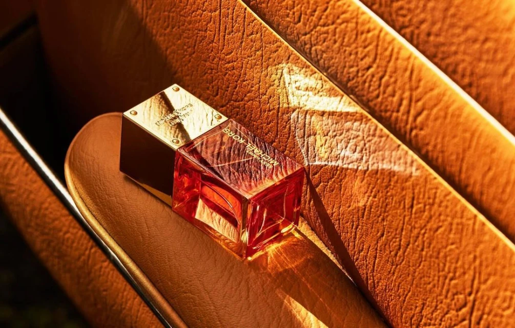 Close-up of a Michael Kors perfume bottle with a gold cap on a textured orange surface, highlighted by a beam of light.
