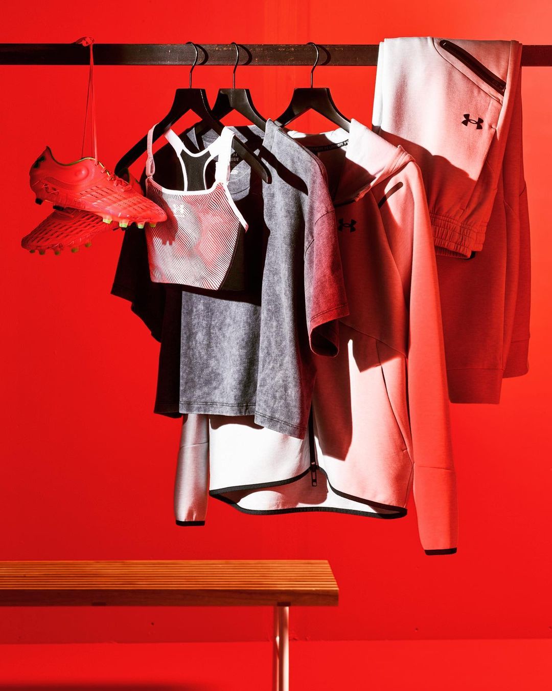 Sports apparel and red cleats hang on a rack against a vibrant red wall, complemented by a wooden bench, creating a dynamic visual.