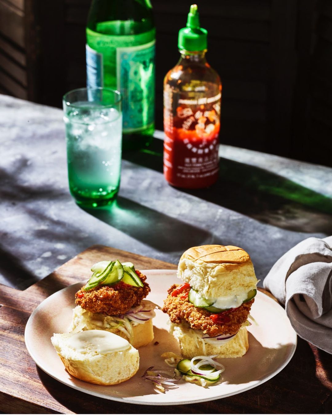 Close-up of two chicken sliders on a plate, garnished with cucumber, served with a bottle of green soda and hot sauce.
