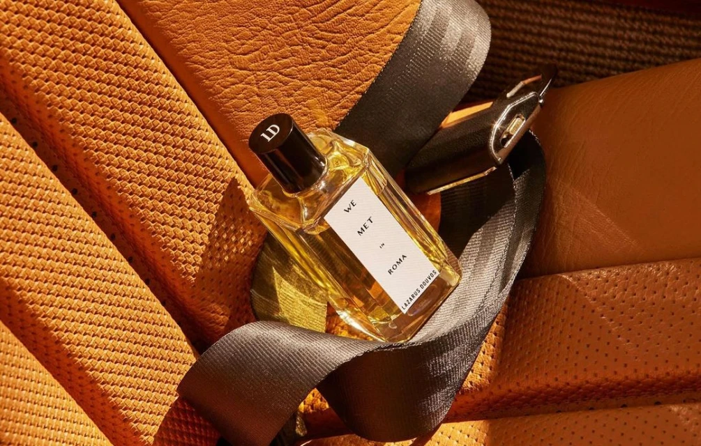 Close-up of a rectangular glass perfume bottle with a black lid, featuring an amber liquid. The bottle is held in place with a car seatbelt.