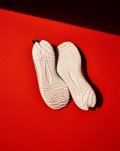 Elevated studio shot captures the textured soles of white Under Armour athletic shoes against a vibrant red background with shadow play.