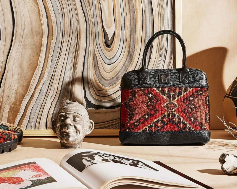 Fashionable handbag with geometric woven pattern, placed alongside open book and sculpture on a table, showcasing artistry and style.