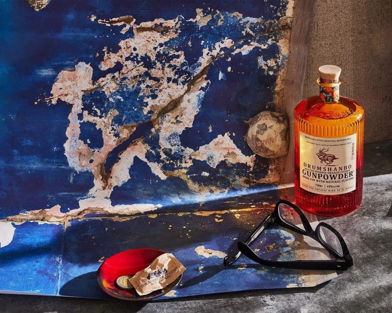 Still life of a Drumshanbo Gunpowder Irish Gin bottle, eyeglasses, dish, and crumpled paper set against a textured blue background.