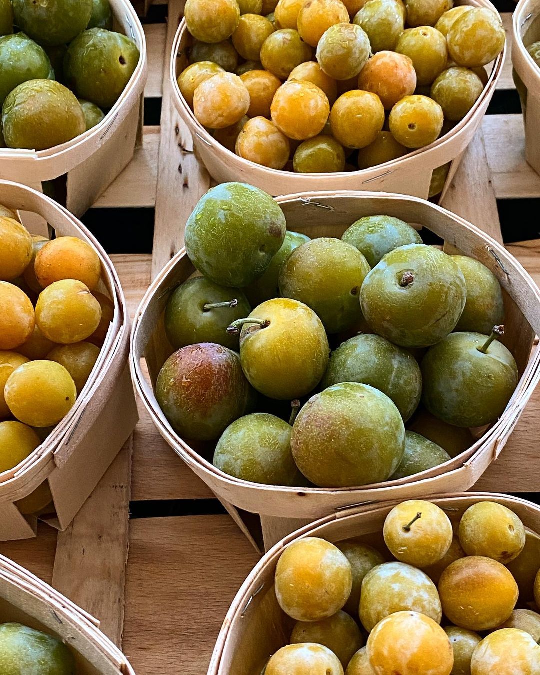 Close-up of several wooden baskets filled with green and yellow plums, showcasing the natural textures and colors of the ripe fruit.