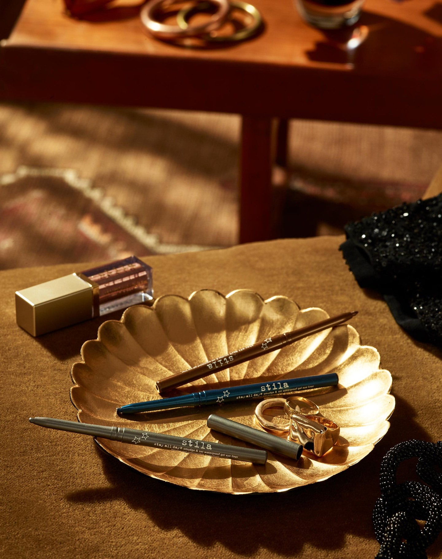 Close-up of gold cosmetic products with Stila eyeliner pencils on a gold plate with jewelry and dark fabric in a warm-toned scene.