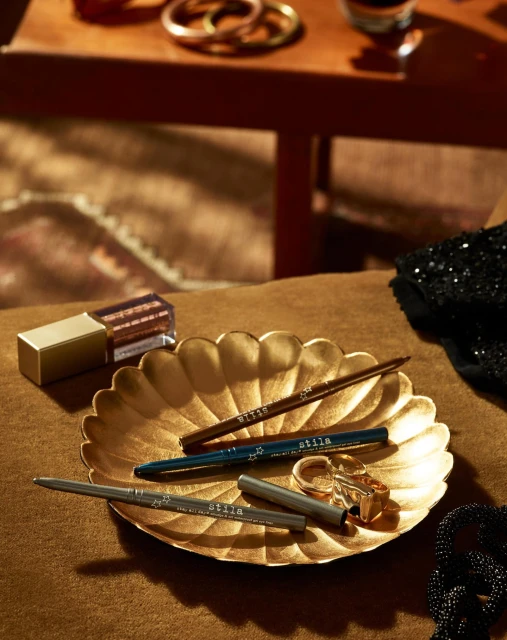 Close-up of gold cosmetic products with Stila eyeliner pencils on a gold plate with jewelry and dark fabric in a warm-toned scene.
