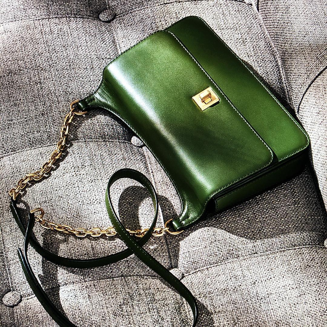 Olive green leather shoulder bag with gold chain strap on a gray textured sofa, in a product studio shot.