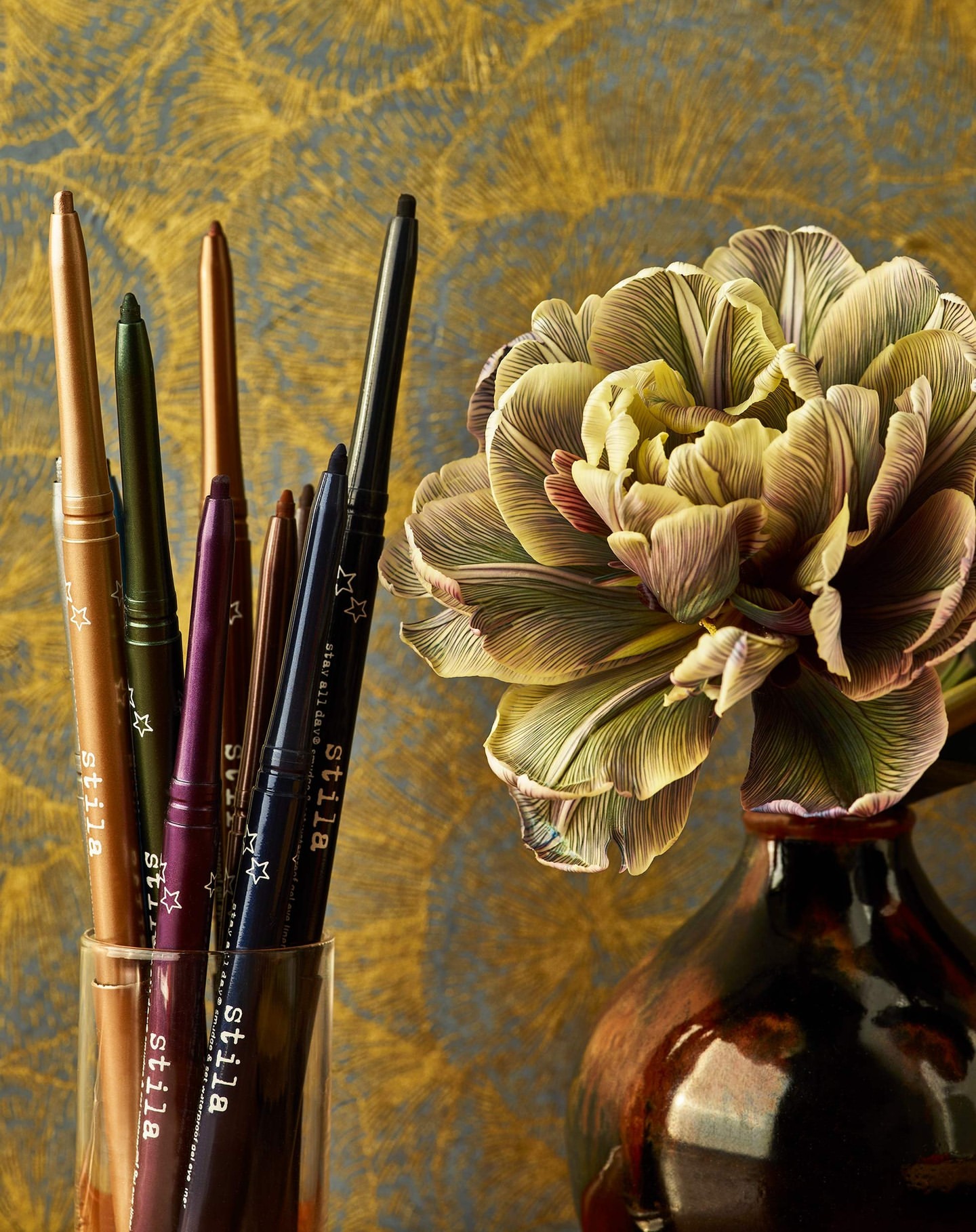 Several Stila eyeliner pencils arranged in a glass with a cream and green tulip in a brown vase against a gold patterned wall.