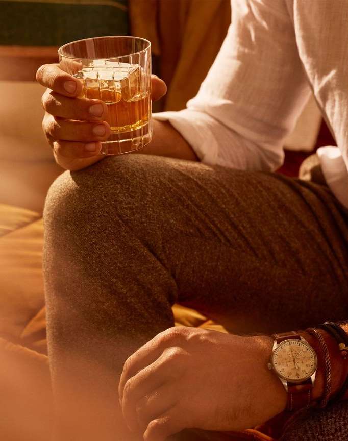 Close-up of a man holding a glass of amber liquid with ice, wearing a white shirt, brown pants, and a wristwatch on his left wrist.