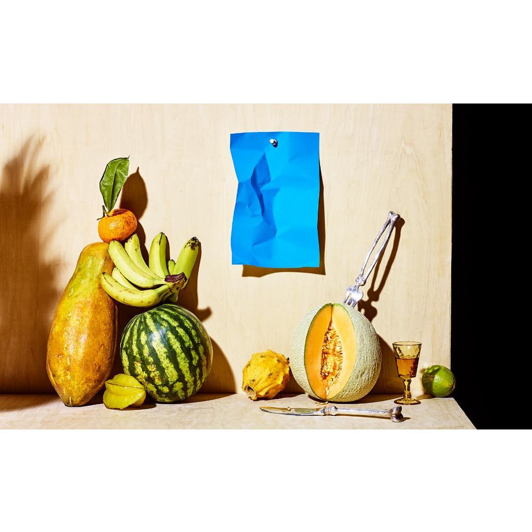 Still life of tropical fruit including a halved cantaloupe, watermelon, papaya, bananas, starfruit, and citrus on a counter with a blue crumpled paper.