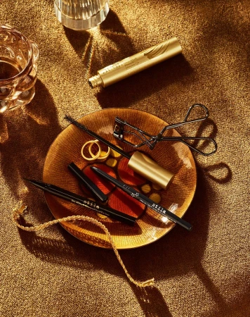 Still life of golden-toned beauty products on a plate, including mascara, eyeliner, an eyelash curler, and gold jewelry.