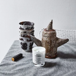 Still life photography of a ceramic teapot, a stack of patterned cups, a scented candle, and a lighter on a grey fabric table.