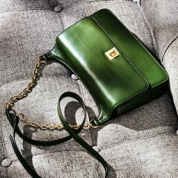 An emerald green leather shoulder bag with a gold clasp and chain strap resting on a textured grey fabric surface.