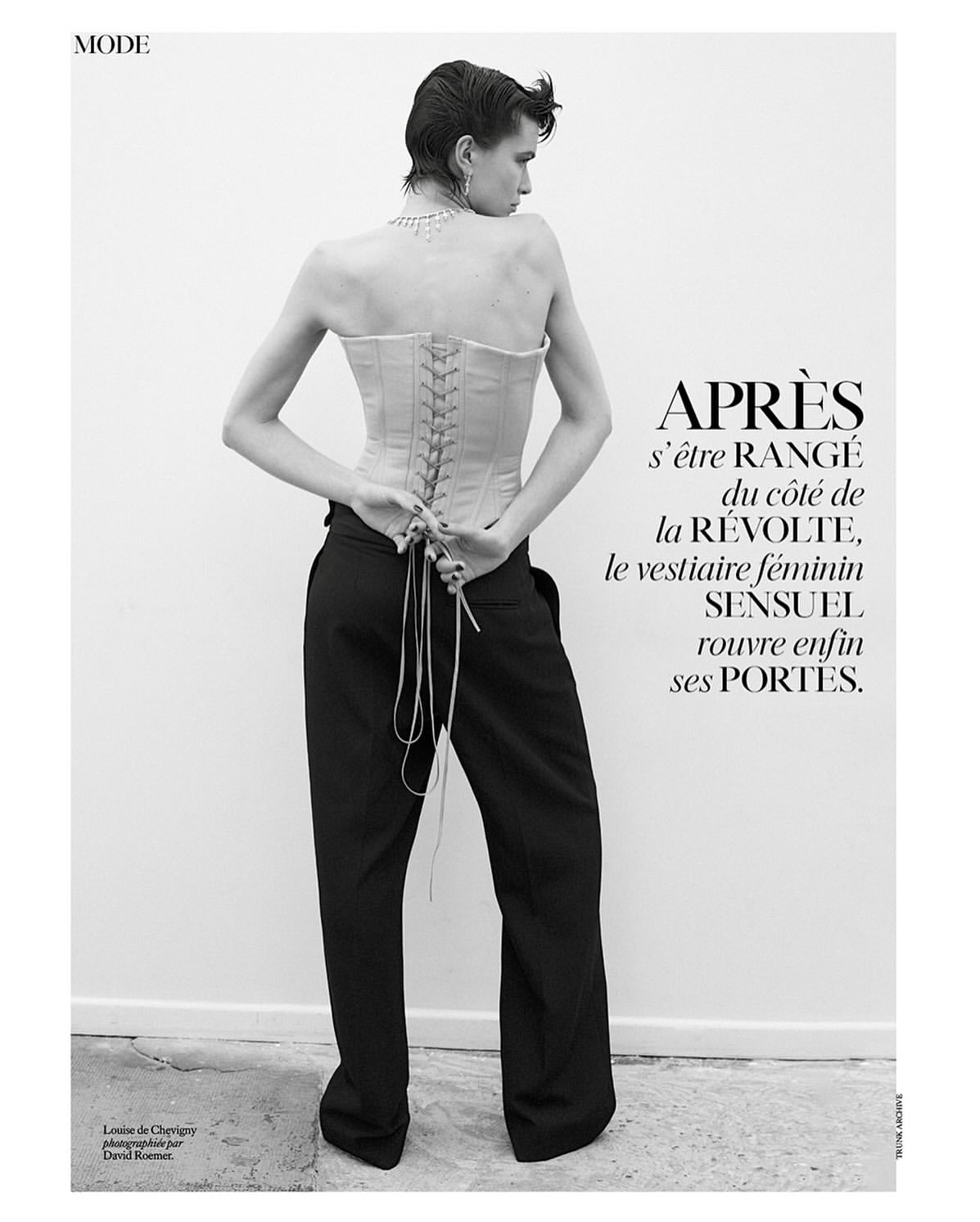 Model with short dark hair wearing a lace-up corset top and black trousers against a plain white wall