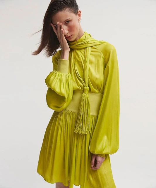 Fashion model wearing a chartreuse silk dress with long sleeves and a tassel detail poses in a studio against a neutral background.