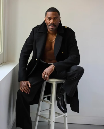 A Black man wearing a black coat and trousers sits on a white stool in a studio in Paris.