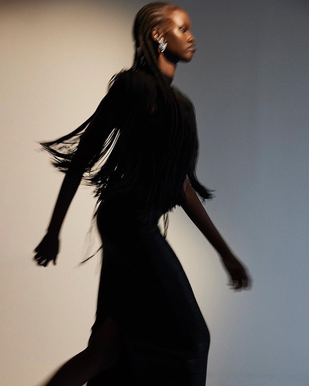 A slender woman in motion wearing a fringe-detailed black dress and large earrings against a soft, gradated background, fashion photography.